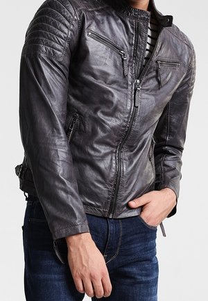 Hombre que lleva una chaqueta de cuero gris oscuro con cremallera, paneles acolchados en los hombros y múltiples bolsillos con cremallera, combinado con vaqueros azul oscuro.