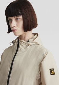 Giacca impermeabile beige chiaro con colletto alto, cerniera frontale e cappuccio regolabile con cordino. Presenta un patch con logo sulla manica sinistra.