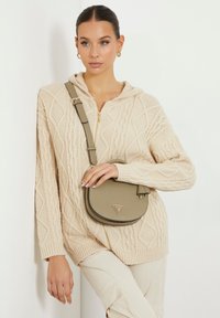 Maglione beige a maglia rasata con cappuccio, caratterizzato da polsini e orlo a coste. Una piccola borsa rotonda di colore verde chiaro con un dettaglio del logo è appoggiata trasversalmente sul corpo.
