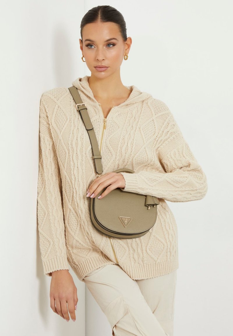 Maglione beige a maglia rasata con cappuccio, caratterizzato da polsini e orlo a coste. Una piccola borsa rotonda di colore verde chiaro con un dettaglio del logo è appoggiata trasversalmente sul corpo.