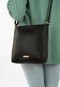 Black leather crossbody bag with pebble texture, side stitching, and gold-tone hardware, worn over a green garment.