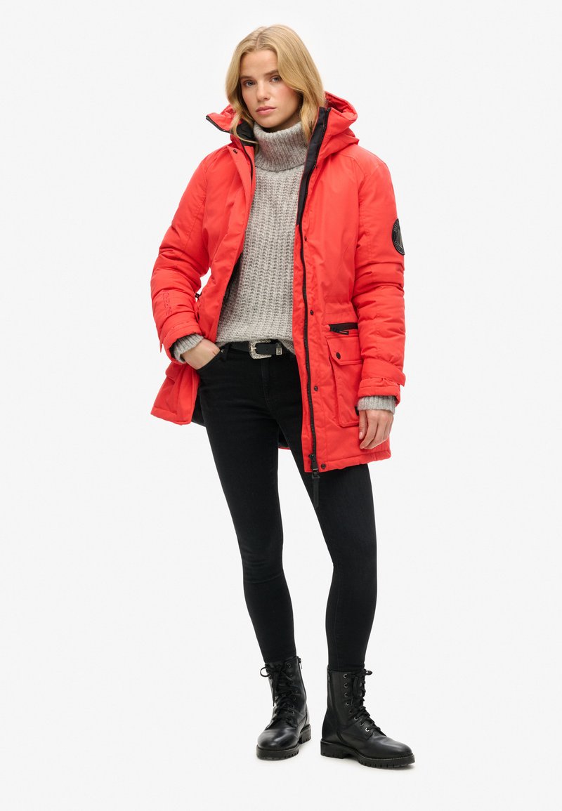 Rote isolierte Parka mit Kapuze, ausgestattet mit einem schwarzen Reißverschluss und Taschen. Getragen über einem grauen Pullover und kombiniert mit engen schwarzen Jeans und Stiefeln.