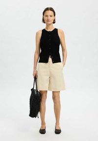 Top senza maniche nero a coste con bottoni, pantaloni corti beige e borsa a rete nera, abbinati a scarpe slip-on nere texturizzate.