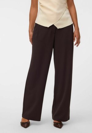 Vrouw draagt wijde zwarte broek, crèmekleurige mouwloze overslagtop en bruine puntige platte schoenen met strikdetails.