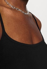 Svart linne med tunna spännen, med en rundad halsringning. Bärs med ett silverkedjesmycke som framhäver en jämn textur.