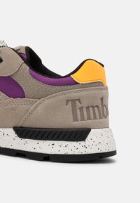 Talons de baskets en daim beige, mesh violet, accent orange, semelle noire et blanche tachetée, avec logo Timberland en relief sur le côté.