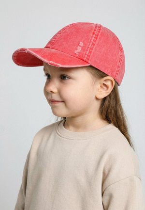 Junges Mädchen mit langen Haaren, das eine verwaschene rote Baseballkappe und ein beiges Sweatshirt trägt, leicht nach links blickend mit einem sanften Lächeln.