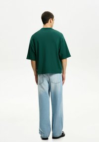 Grön bomullspolo med avslappnad passform, korta ärmar och krage, tillsammans med ljusblå vida jeans och svarta skor.