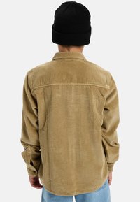 Person in einer beigen Cordjacke, hellblauen Jeans und einer schwarzen Strickmütze, der mit dem Rücken zu einer schlichten weißen Hintergrunder steht.