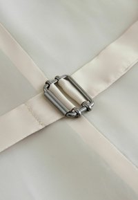 Nahaufnahme eines beigen Stoffbands, das durch eine rechteckige Metallschnalle auf glattem, hellfarbenem Material befestigt ist.