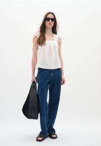 Haut sans manches blanc avec un design texturé, jeans bleus taille haute, et sac en cuir noir, associé à des lunettes de soleil noires et des sandales.