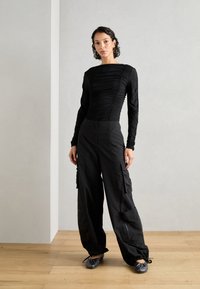 Top negro de manga larga con textura fruncida, combinado con pantalones cargo negros holgados. El modelo se encuentra de pie sobre un piso de madera frente a un fondo gris claro.