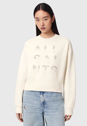 Młoda kobieta z długimi ciemnymi włosami nosi białą bluzę z napisem "ALL SAINTS" oraz jasnoniebieskie jeansy z przetarciami.