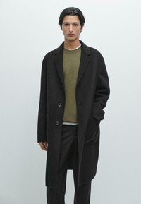 Cappotto lungo nero da uomo con revers a punta, dotato di due bottoni, tasche e un tessuto in lana texture, indossato sopra un maglione verde.