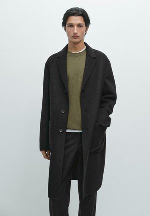 Cappotto lungo nero da uomo con revers a punta, dotato di due bottoni, tasche e un tessuto in lana texture, indossato sopra un maglione verde.