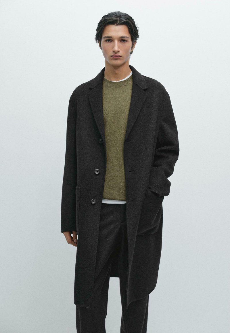 Langer schwarzer Herrenmantel mit Reverskragen, ausgestattet mit zwei Knöpfen, Taschen und einem strukturierten Wollstoff, getragen über einem grünen Pullover.