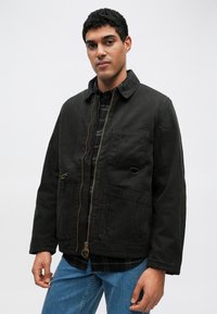 Czarna kurtka na płótnie zapinana na zamek z dwoma przednimi kieszeniami i kołnierzem. Nosi się ją na koszuli w kratę, z widocznymi dżinsami.