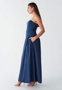 Námornícky modré priliehavé šaty bez ramienok s nabrúsenou sukňou, bočnými vreckami a hladkým materiálom, v kombinácii so svetlými vysokými sandálmi na podpätku.