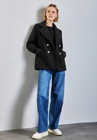 Abrigo de doble botonadura negro con grandes botones plateados, combinado con pantalones de mezclilla de tiro alto y pernera ancha en azul claro. Textura suave, diseño a medida.