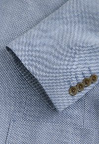 Svetlomodrá texturovaná látka s vzorom k továči, so zrolovaným rukávom a štyrmi hnedými gombíkmi s jemným leskom.