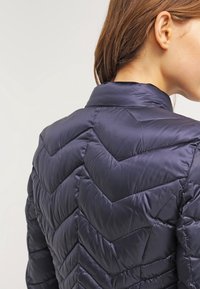 Doudoune bleu marine avec un design matelassé en chevrons, col montant et texture de tissu lisse et brillante. Vue arrière affichée.