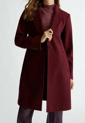 Frau trägt einen langen bordeauxfarbenen Mantel über einem passenden gerippten Pullover und hält mit einer Hand das Revers des Mantels.