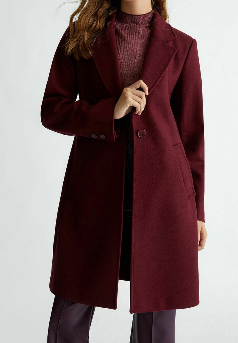 Femme portant un long manteau bordeaux sur un pull côtelé assorti, tenant le revers du manteau d'une main.