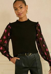 Fekete felső fodros nyakkivágással, áttetsző hosszú ujjakkal, amelyek rózsaszín szívmintákkal díszítettek, magas derekú fekete nadrággal és arany övcsattal párosítva.