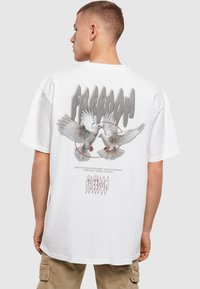 Hvid oversized T-shirt med et grafisk motiv af to duer i flugt, omgivet af ordet "FRIHED" i fede, stiliserede bogstaver.