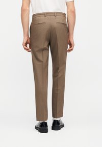 Άνδρας που φοράει καφέ ραμμένο παντελόνι, λευκές κάλτσες και μαύρα επίσημα παπούτσια, στέκεται με πλάτη σε λευκό φόντο.