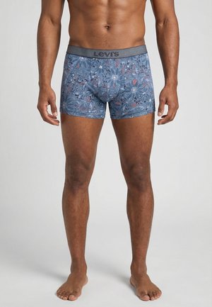 Modelo masculino con bóxers Levi's azules con estampado floral y cintura gris, de pie descalzo sobre un fondo gris liso.