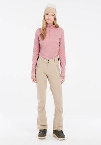 Leichtes, rosa Langarmoberteil mit einem Viertelreißverschluss, kombiniert mit braunen Hosen mit seitlichen Reißverschlüssen und hoher Taille, ergänzt durch dunkle Stiefel.