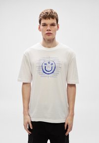 Camiseta blanca de algodón con un diseño gráfico azul que incluye el texto "HGO" y un contorno de cara sonriente, mangas cortas y un ajuste holgado.