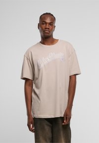 Beige oversized t-skjorte i bomull med et lilla trykk "Urban Classics 05" på brystet. Enkel rund halsutskjæring og korte ermer.