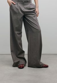 Grijze wijde broek met voorplooien; heeft een soepele textuur en een op maat gemaakte pasvorm. Donkerburgundy schoenen met gouden studs maken de look compleet.