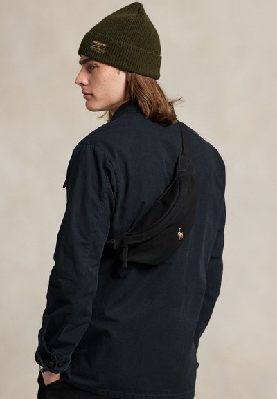 Młody mężczyzna w oliwkowej zielonej czapce, ciemnej koszuli i małej czarnej torbie na pasku przez ramię, zwrócony w lewo na neutralnym beżowym tle.