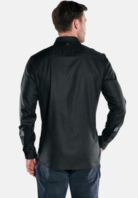 Ein Mann, der von hinten zu sehen ist, trägt ein schwarzes langärmeliges Hemd mit Knöpfen und dunkelblaue Jeans vor einem schlichten hellen Hintergrund.