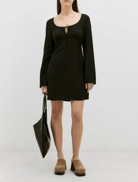 Zwarte lange jurk met lange mouwen en een ronde halslijn, van textuurstof, met een strikdetail op de borst, gecombineerd met een zwarte tote en bruine klompen.