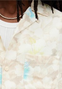 Primo piano di una persona che indossa una camicia a fiori crema sopra una maglia bianca lavorata a maglia, con una collana di perline argentate e dreadlocks.