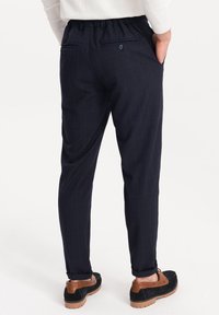 Marineblauwe geruite broek met een tapered fit, voorzien van een elastische tailleband en achterzakken met knoopsluitingen. Paar donkere schoenen zichtbaar.