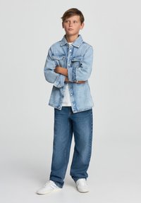 Jasnoniebieska kurtka dżinsowa z zapięciem na guziki, dwoma kieszeniami na piersi, biała koszulka pod spodem, w zestawieniu z luźnymi niebieskimi dżinsami i białymi trampkami.