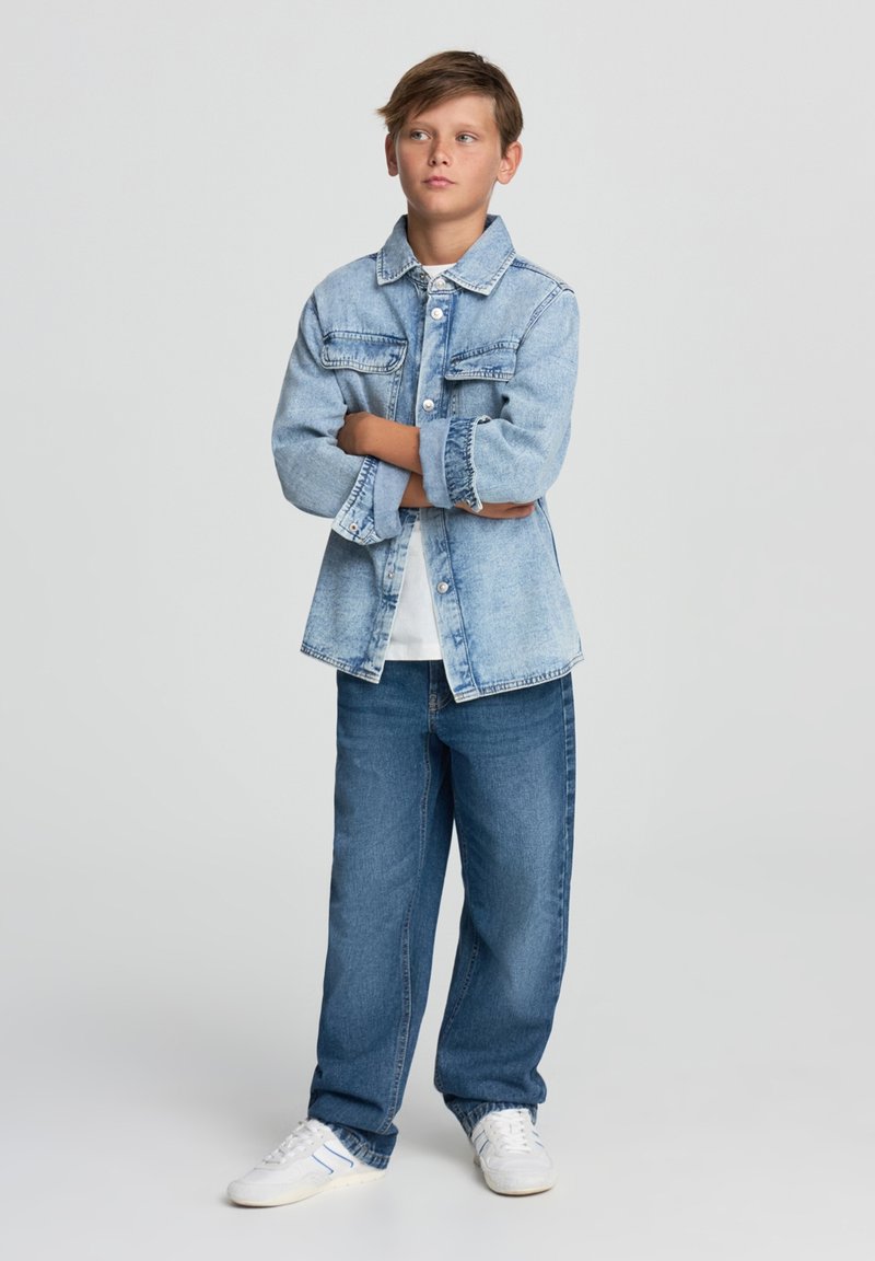 Jasnoniebieska kurtka dżinsowa z zapięciem na guziki, dwoma kieszeniami na piersi, biała koszulka pod spodem, w zestawieniu z luźnymi niebieskimi dżinsami i białymi trampkami.