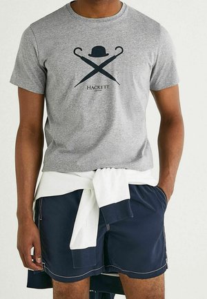 T-shirt en coton gris avec un motif graphique noir représentant des parapluies croisés et un chapeau melon, associé à un short bleu marine et un pull blanc noué à la taille.
