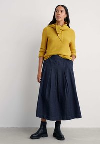 Geel geribbelde trui met een gestrikte kraag, gecombineerd met een marineblauwe geplooide denimrok. Zwarte enkellaarzen maken de outfit compleet. Gladde textuur.