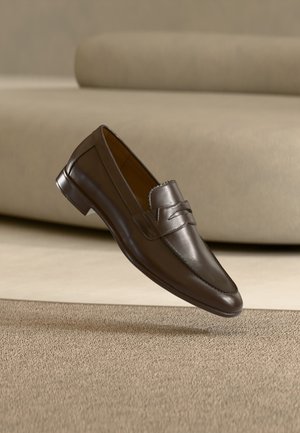 Mocassin en cuir marron foncé flottant au-dessus d'un tapis beige avec un canapé courbé beige en arrière-plan.