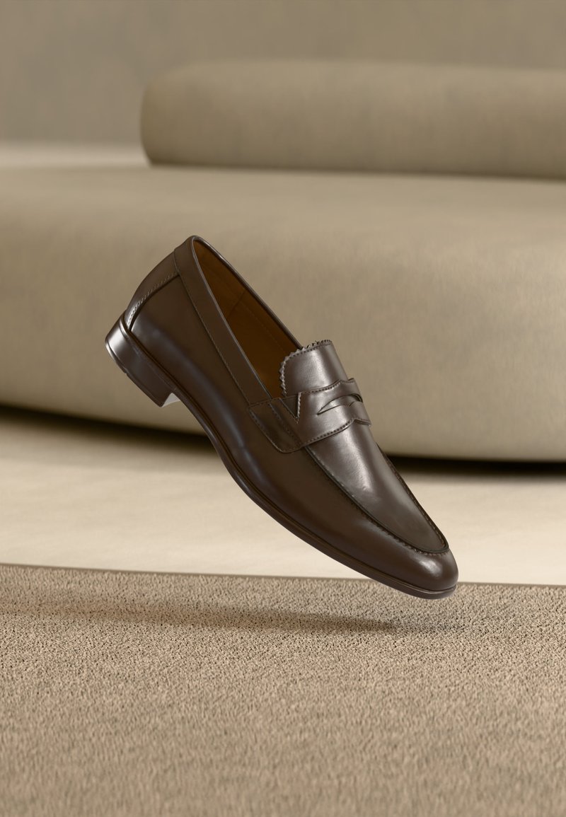 Mocassin en cuir marron foncé flottant au-dessus d'un tapis beige avec un canapé courbé beige en arrière-plan.