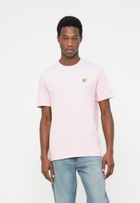 Tricou roz deschis din bumbac, cu mâneci scurte și decolteu rotund. Prezintă un mic logo galben în formă de fluture pe partea stângă a pieptului.