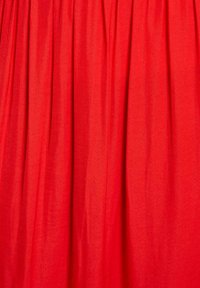 Tessuto rosso con una texture liscia, caratterizzato da pieghe verticali che creano una silhouette fluida. La luce si riflette dolcemente sulla superficie.