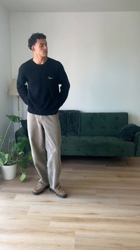 Suéter preto com textura canelada, calças de camurça bege e sapatos estampados. A pé à frente de um sofá verde, com uma planta e um chão de madeira.