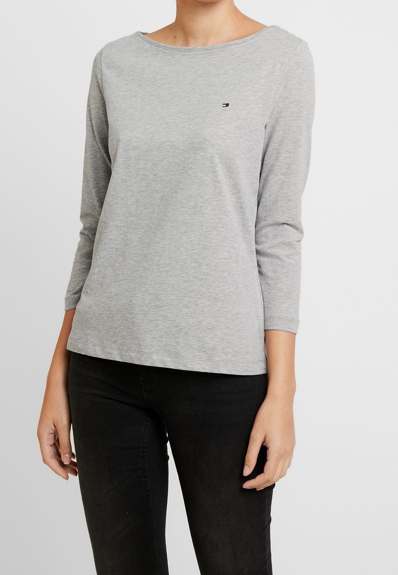 Graues Langarmshirt aus weichem Stoff, mit rundem Halsauschnitt und kleinem Logo auf der Brust. Kombiniert mit schwarzen Jeans.